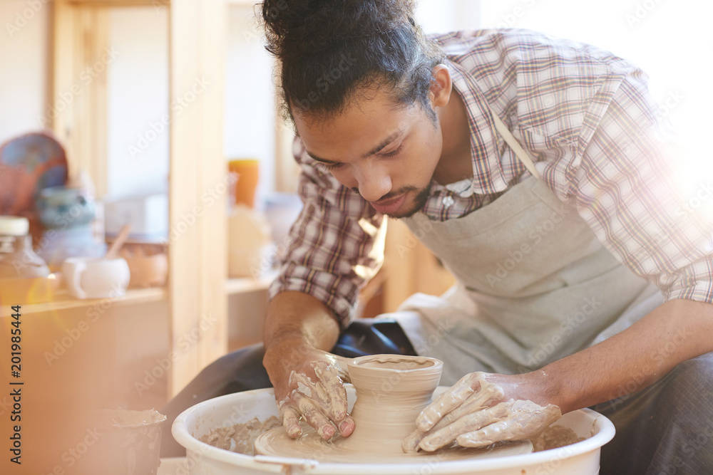 Portrait of young handsome mixed race male potter making clay vase on spinning pottery wheel in workshop Portrait of young handsome mixed race male potter making clay vase on spinning pottery wheel in workshop