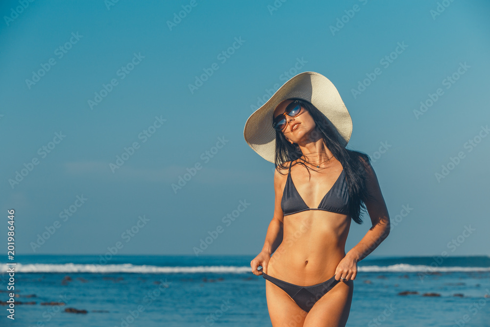 Beach vacation. Hot beautiful woman in sunhat and bikini standing with her arms raised to her head enjoying looking view of beach ocean on hot summer day. Photo from Hapuna beach, Big Island, Hawaii.