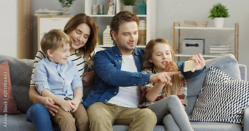 Caucasian happy family sitting on the sofa in the living room, hugging while the father showing something on the smartphone and they discussing it emotionally. At home. Indoor Caucasian happy family sitting on the sofa in the living room, hugging while the father showing something on the smartphone and they discussing it emotionally. At home. Indoor