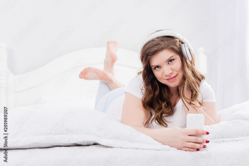 portrait of young woman in pajama and headphones with smartphone lying on bed at home portrait of young woman in pajama and headphones with smartphone lying on bed at home