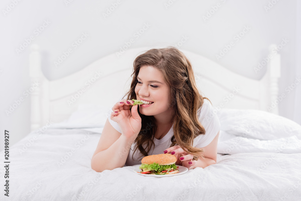 overweight woman in pajama eating burger on bed at home overweight woman in pajama eating burger on bed at home