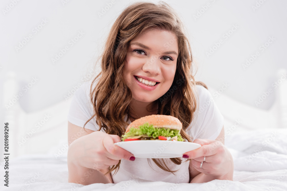 portrait of overweight smiling woman in pajama with burger on bed at home portrait of overweight smiling woman in pajama with burger on bed at home
