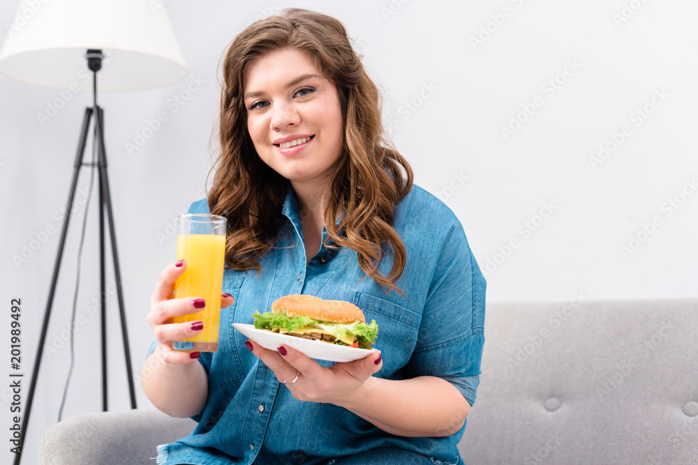 overweight smiling woman with glass of juice and burger on plate in hands at home overweight smiling woman with glass of juice and burger on plate in hands at home