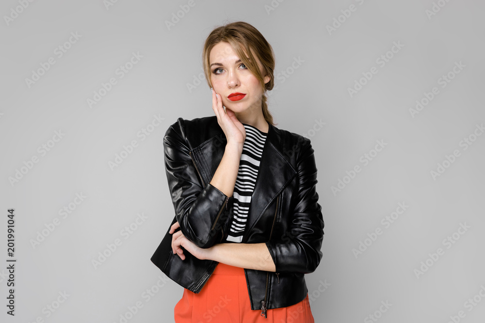Attractive young serious blonde girl in striped blouse and leather jacket thinking waiting on gray background Attractive young serious blonde girl in striped blouse and leather jacket thinking waiting on gray background
