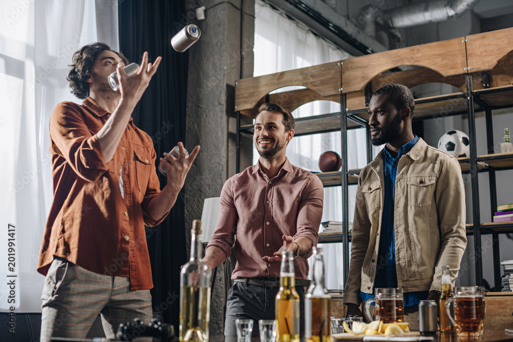 low angle view of multiethnic men looking at friend juggling with beer cans low angle view of multiethnic men looking at friend juggling with beer cans