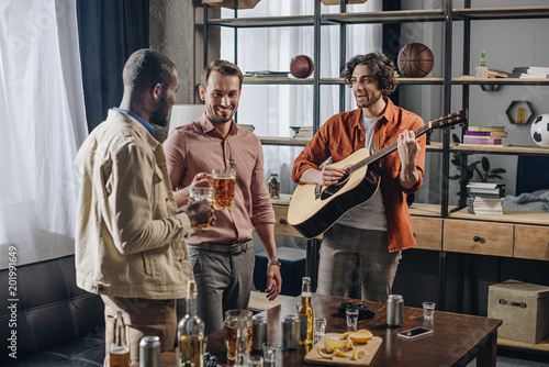 happy multiethnic male friends drinking beer and playing guitar together