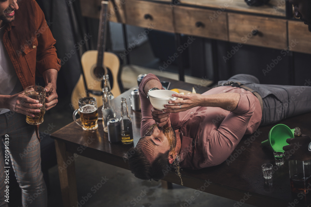 young man lying on table and drinking beer from funnel while friends looking at him