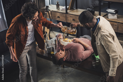 multiethnic friends pouring beer in funnel and man drinking while lying on table