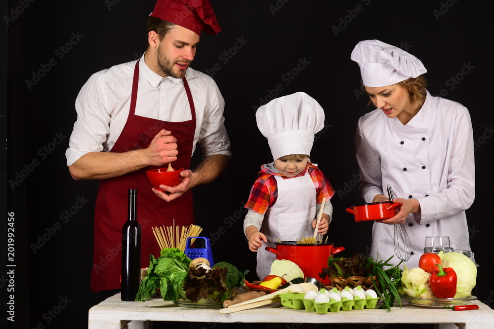 Child with parents cooking at kitchen table. Adorable kid in chef hat. Happy loving family preparing dinner together. Homemade food. Child with parents cooking at kitchen table. Adorable kid in chef hat. Happy loving family preparing dinner together. Homemade food.
