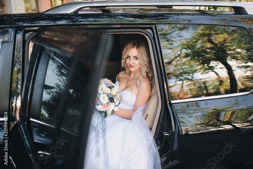 A beautiful bride with curly hair is sitting in the car and looking out the window. A nice bride in the city comes out of the car.