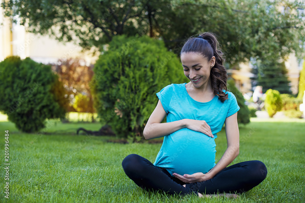Rest for pregnant mothers. Beautiful young pregnant woman in sportswear sitting on grass in park in lotus pose doing breathing exercises Rest for pregnant mothers. Beautiful young pregnant woman in sportswear sitting on grass in park in lotus pose doing breathing exercises