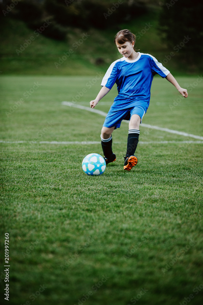 Teen girl playing soccer at local stadium outside on grass field kicking the ball. Children playing football, favourite sport, football fever worldwide. Teen girl playing soccer at local stadium outside on grass field kicking the ball. Children playing football, favourite sport, football fever worldwide.
