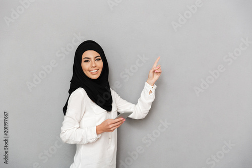 Portrait of gorgeous arab woman in headscarf with oriental makeup holding silver smartphone and pointing finger at copy space, isolated over gray background