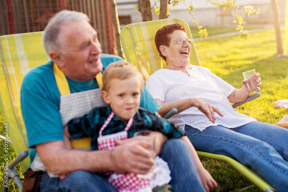 Elderly man is sitting and holding his little adorable toddler grandson his wife is sitting beside him and laughing.