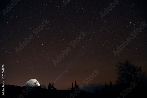 tourist with flashlight near his camp tent under a sky full of stars, night mountain loneliness