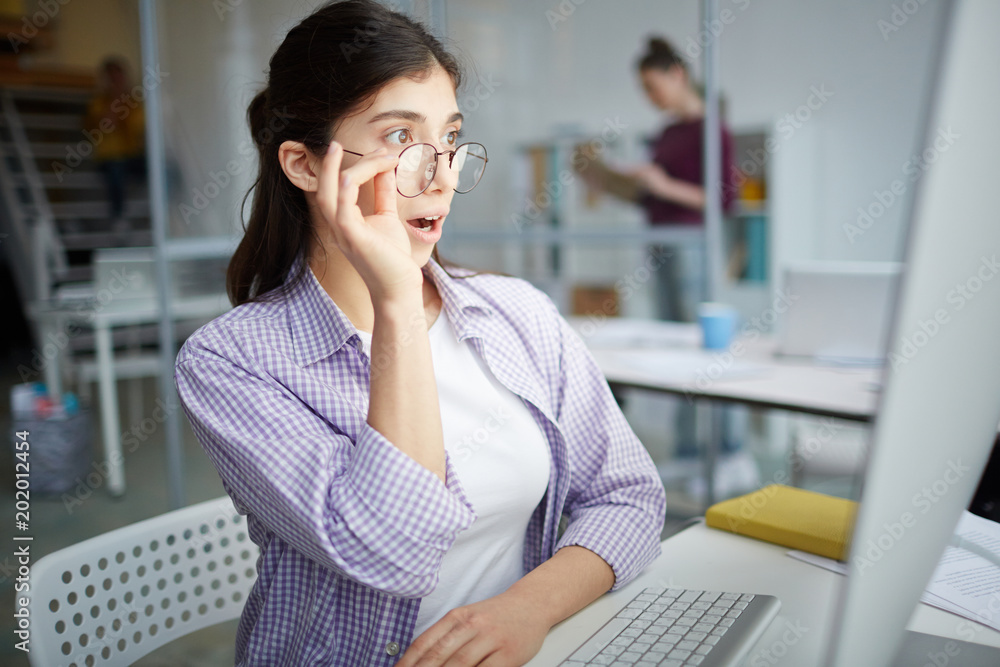 Businesswoman in eyeglasses sitting at the table and looking at monitor with surprise Businesswoman in eyeglasses sitting at the table and looking at monitor with surprise