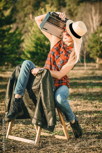 an attractive young hipster girl sits on a chair in the Park, holding a tape recorder on her shoulder, in a straw hat on her head and sunglasses.