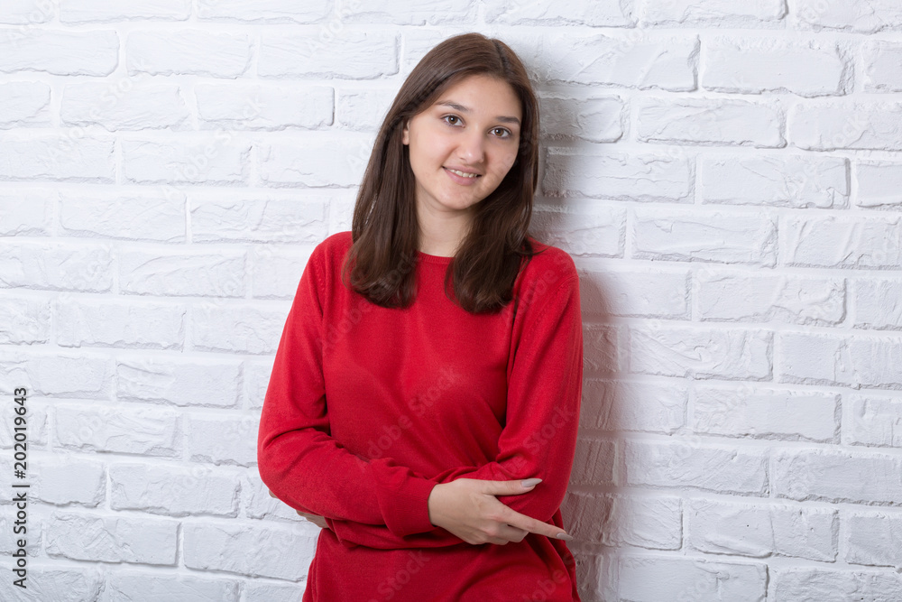 A young pretty smiling girl in a red sweater and a hat against a modern white brick wall in the loft style. She looks at the viewer. Copy space. A young pretty smiling girl in a red sweater and a hat against a modern white brick wall in the loft style. She looks at the viewer. Copy space.