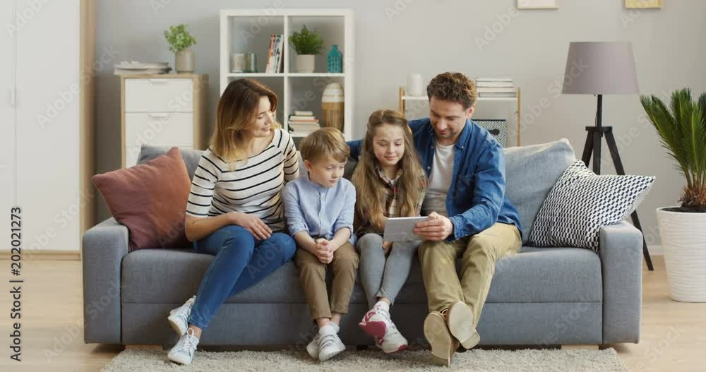Joyful caucasian family with two kids sitting on the sofa, smiling while watching something on the tablet computer. At home. Indoors Joyful caucasian family with two kids sitting on the sofa, smiling while watching something on the tablet computer. At home. Indoors