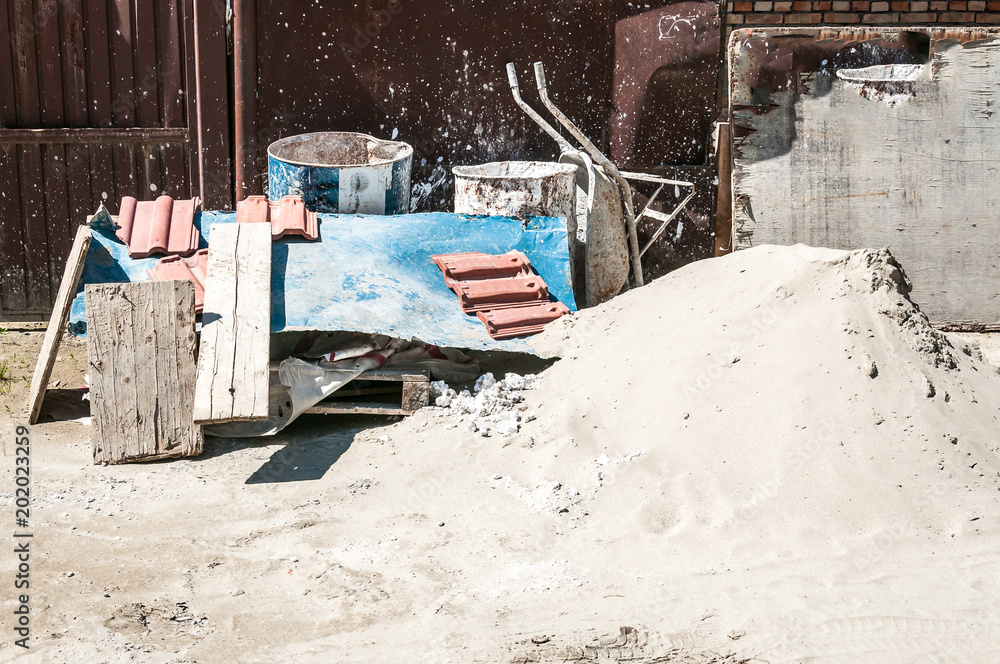 Pile of sand with trolley and other architectural material and tools in front of the residential building constriction site on the street