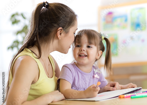 Mother whispering her child daughter while she draws a picture in nursery