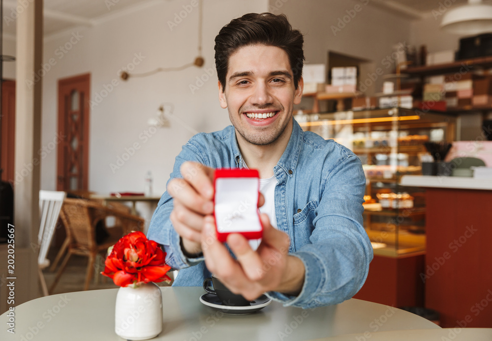 Attractive smiling man sitting at table in restaurant and proposing to his girlfriend, with giving box with engagement ring at camera