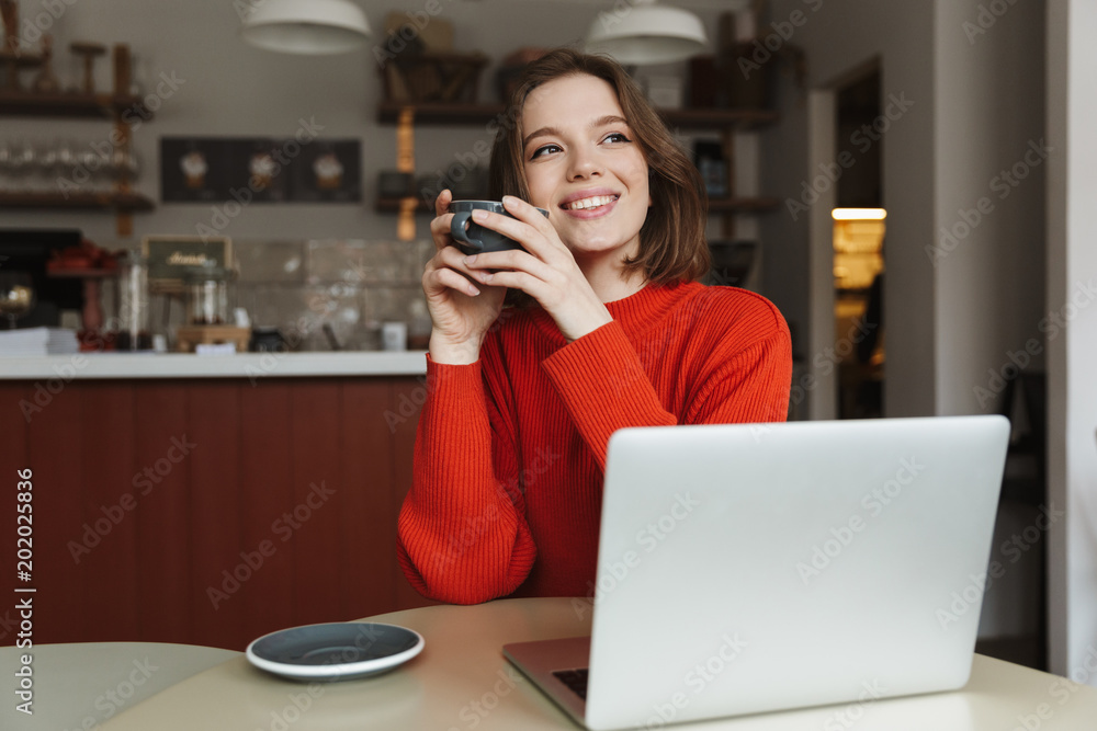 Image of happy caucasian woman 20s smiling, while using laptop and drinking coffee in cafe Image of happy caucasian woman 20s smiling, while using laptop and drinking coffee in cafe