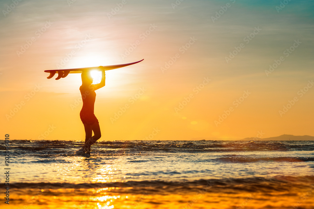 Silhouette of woman holding surfboard turn on the head walking on the edge of sea wave, retuning to home station after surfed in the sea at sunset light