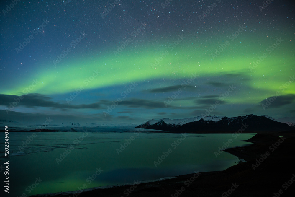 Northern lights aka Aurora Borealis glowing on sky in Jokulsarlon glacier lagoon with snow capped mountains in background at night in Iceland