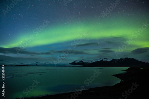 Northern lights aka Aurora Borealis glowing on sky in Jokulsarlon glacier lagoon with snow capped mountains in background at night in Iceland © Jamo Images