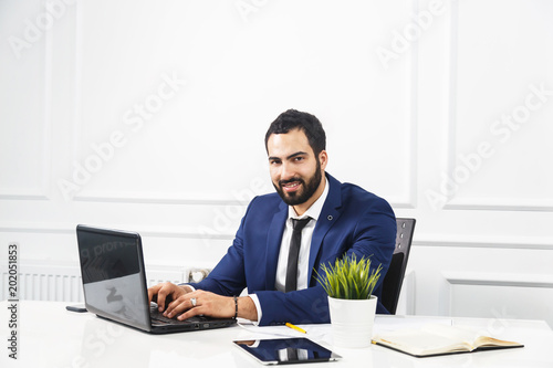 Bearded businessman dressed in blue suit work with laptop in white office interior