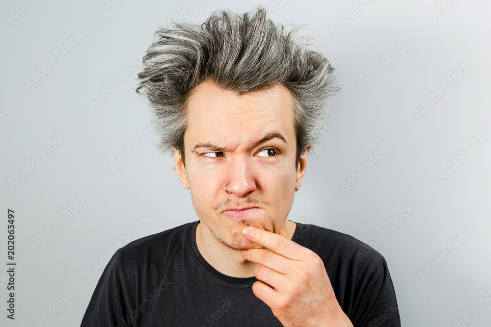 Unshorn and unshaven young guy with piercings on his face, thinking, holding hand at the face and looking aside, on gray background. Unshorn and unshaven young guy with piercings on his face, thinking, holding hand at the face and looking aside, on gray background.