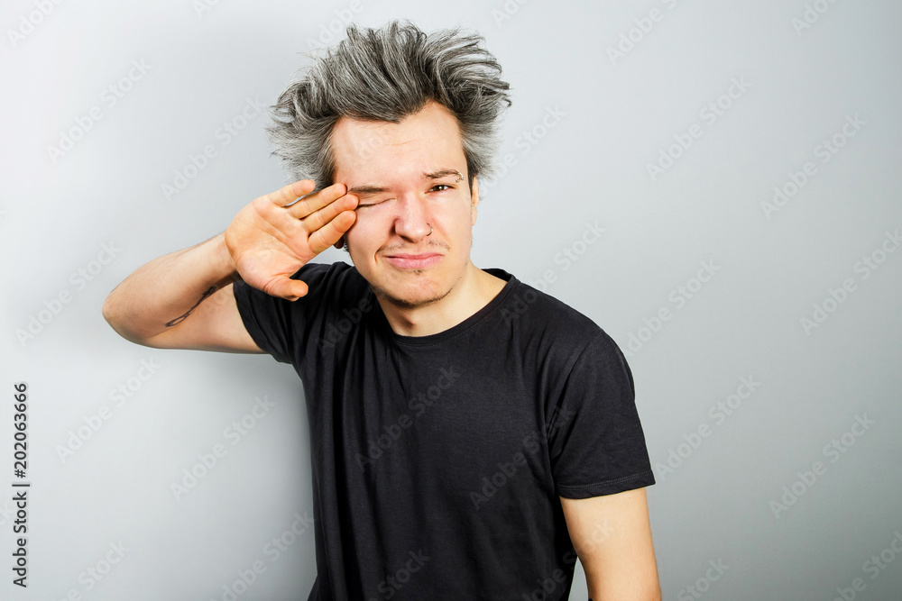 Sleepy, unshorn and unshaven young guy with piercings on his face wipes the eye on gray background. Sleepy, unshorn and unshaven young guy with piercings on his face wipes the eye on gray background.