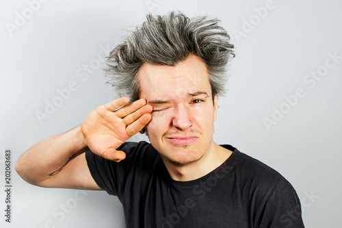 Sleepy, unshorn and unshaven young guy with piercings on his face wipes the eye on gray background. photo