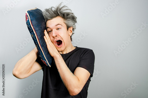 Unshorn and unshaven young guy with piercings on his face sleeps on the pillow and yawn, on gray background. photo