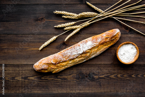 Fresh bread concept. Crispy french baguette near ears of wheat and bowl with flour on dark wooden background top view