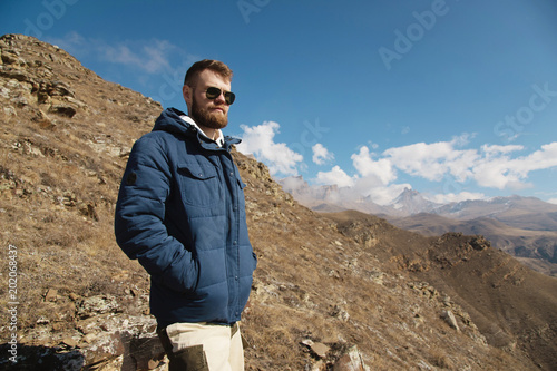 Hipster traveler in a down jacket and sunglasses stands on a mountain slope against the backdrop of epic rocks and smiles. The concept of finding happiness in the mountains