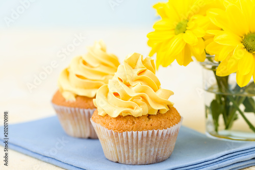 Cupcake with yellow cream decoration on blue napkin and bouquet of yellow chrysanthemum in small glass.