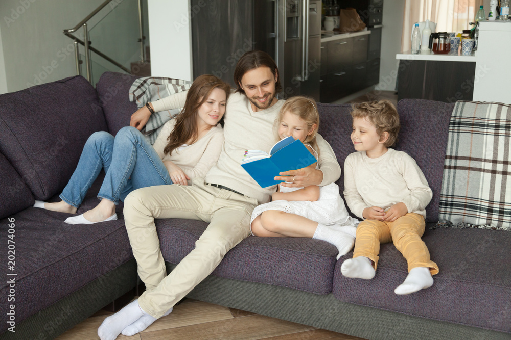 Happy young family with children reading book together sitting on sofa at home, smiling parents relaxing on comfortable couch at home with kids son daughter having fun with storybook in living room Happy young family with children reading book together sitting on sofa at home, smiling parents relaxing on comfortable couch at home with kids son daughter having fun with storybook in living room