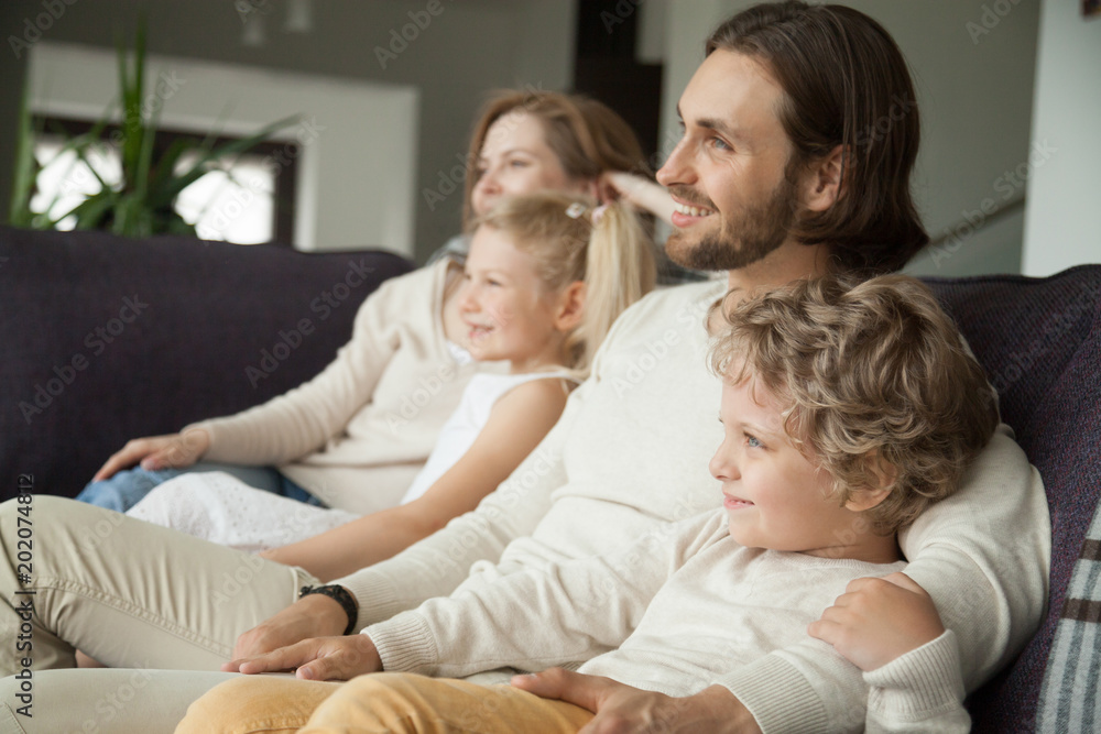 Happy family with children sitting on sofa watching tv, young parents embracing son daughter relaxing on couch together, smiling couple with kids boy and girl having fun on weekend at home, side view