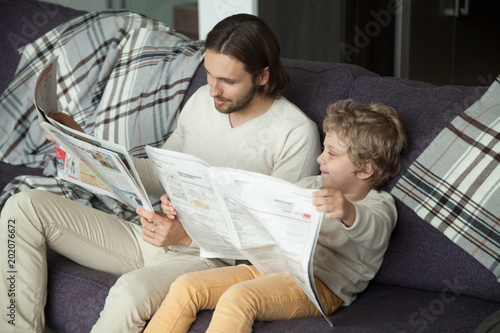 Photos Cute kid son holding newspaper sitting on sofa with dad, curious funny clever bo