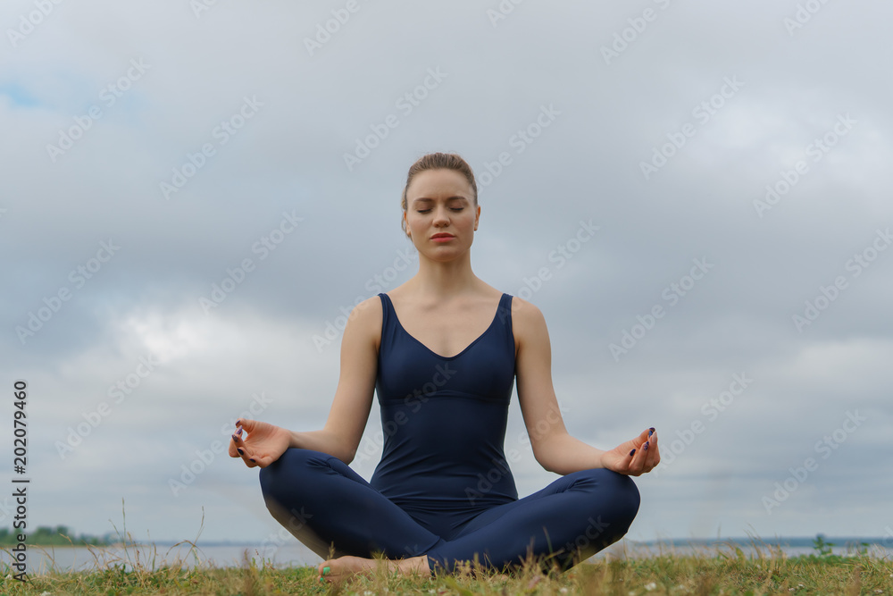 Young girl in blue sportswear sitting in yoga pose, cloudy sky and lake on background.