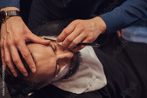 Bearded male sitting in an armchair in a barber shop while hairdresser shaves his beard with a dangerous razor.