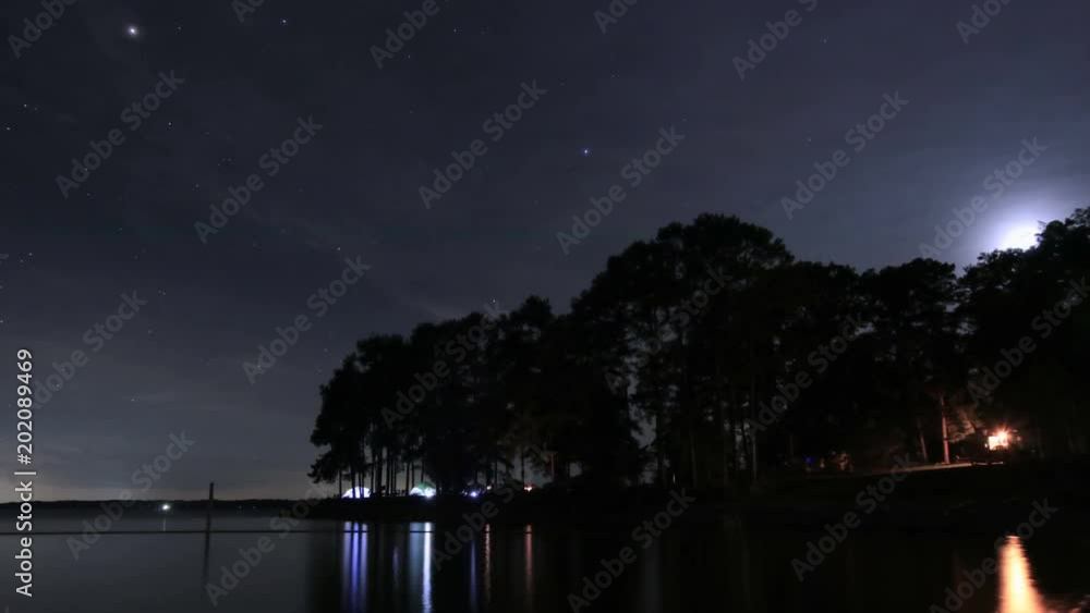 Time lapse of campground across a lake at night with the stars and moon moving as clouds roll in above the trees and glowing tents Time lapse of campground across a lake at night with the stars and moon moving as clouds roll in above the trees and glowing tents