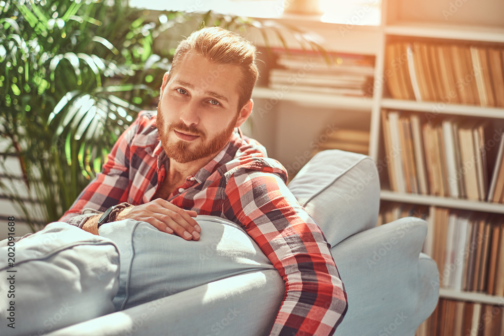 Young handsome redhead model man sits on a sofa in the room with a loft interior. Young handsome redhead model man sits on a sofa in the room with a loft interior.