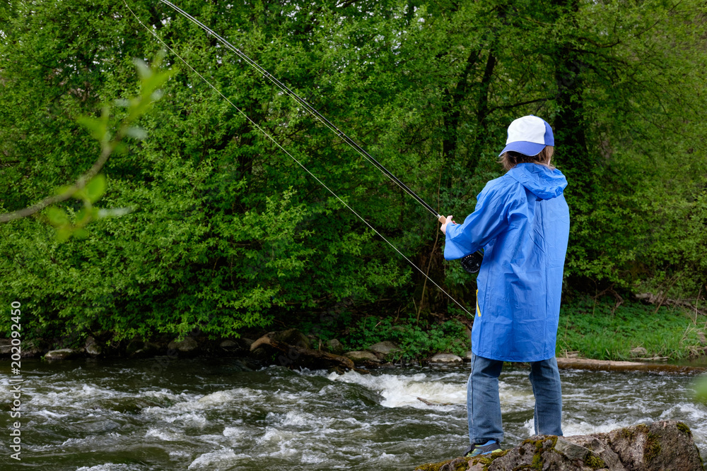 boy, dressed in blue waterproof raincoat and cap, fishing on the mountain streamy river. View from back
