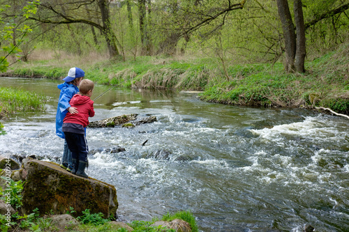 two boys brothers, dressed in red and blue raincoat, are fishing together on the mount river
