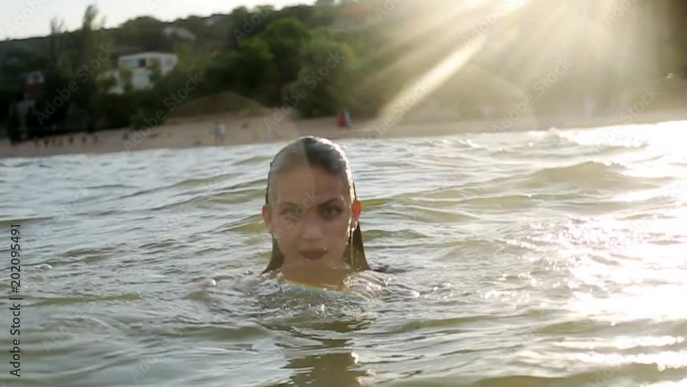 Attractive woman with big breasts emerges from water in a sea water on sunset. Pretty wet smiling girl wearing swimsuit and looks to the camera. Slow motion. Attractive woman with big breasts emerges from water in a sea water on sunset. Pretty wet smiling girl wearing swimsuit and looks to the camera. Slow motion.