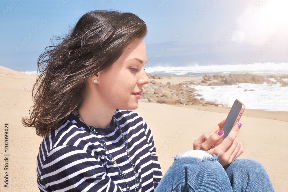 Smiling woman in a striped T-shirt with smartphone relaxing on beach, summer day