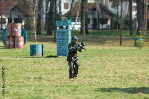 Boy in the camouflage holds a paintball gun in one hand and protective helmet , standing on the field with group of players on the background